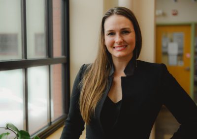 Full Color headshot of Director Julianna Dodson of Radically Rural. She is standing next to a large industrial window in a nondescript commercial building, leaning on the sill and smiling directly into the camera.