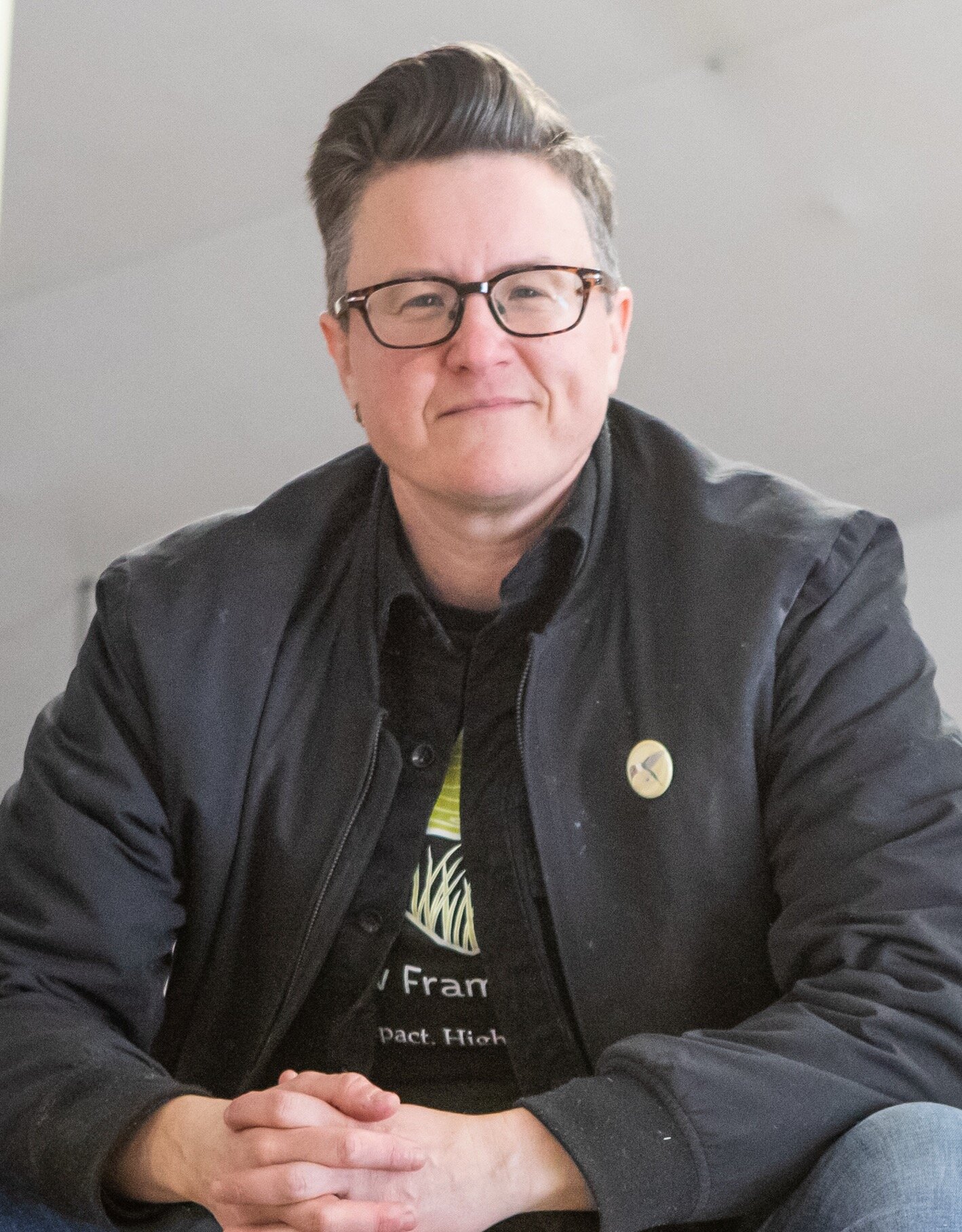 Full Color headshot of Director Julianna Dodson of Radically Rural.  She is standing next to a large industrial window in a nondescript commercial building, leaning on the sill and smiling directly into the camera.